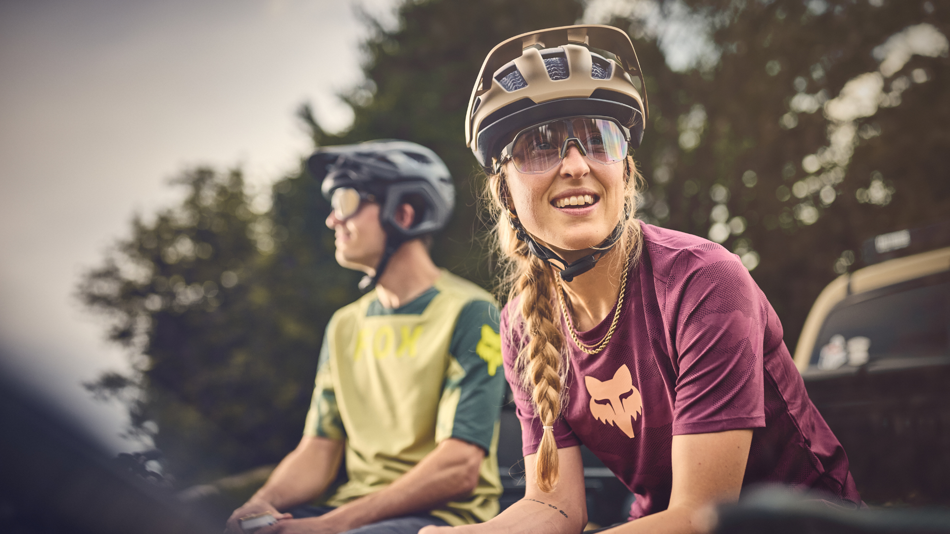 Two people sitting outdoors, one wearing a helmet and sunglasses, with trees in the background.