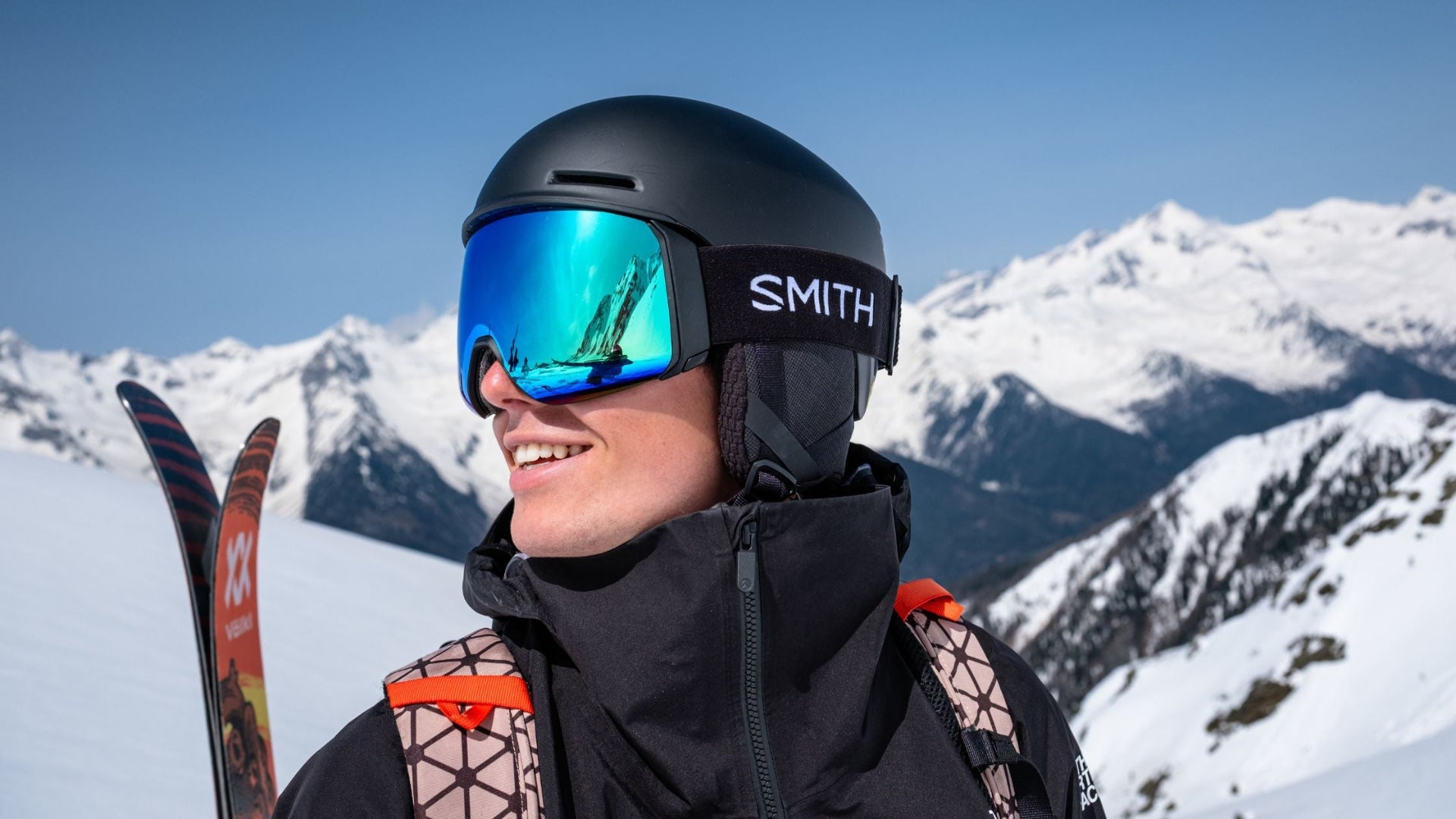 Person wearing a black helmet with 'Smith' logo and blue goggles, holding skis with snowy mountains in the background.