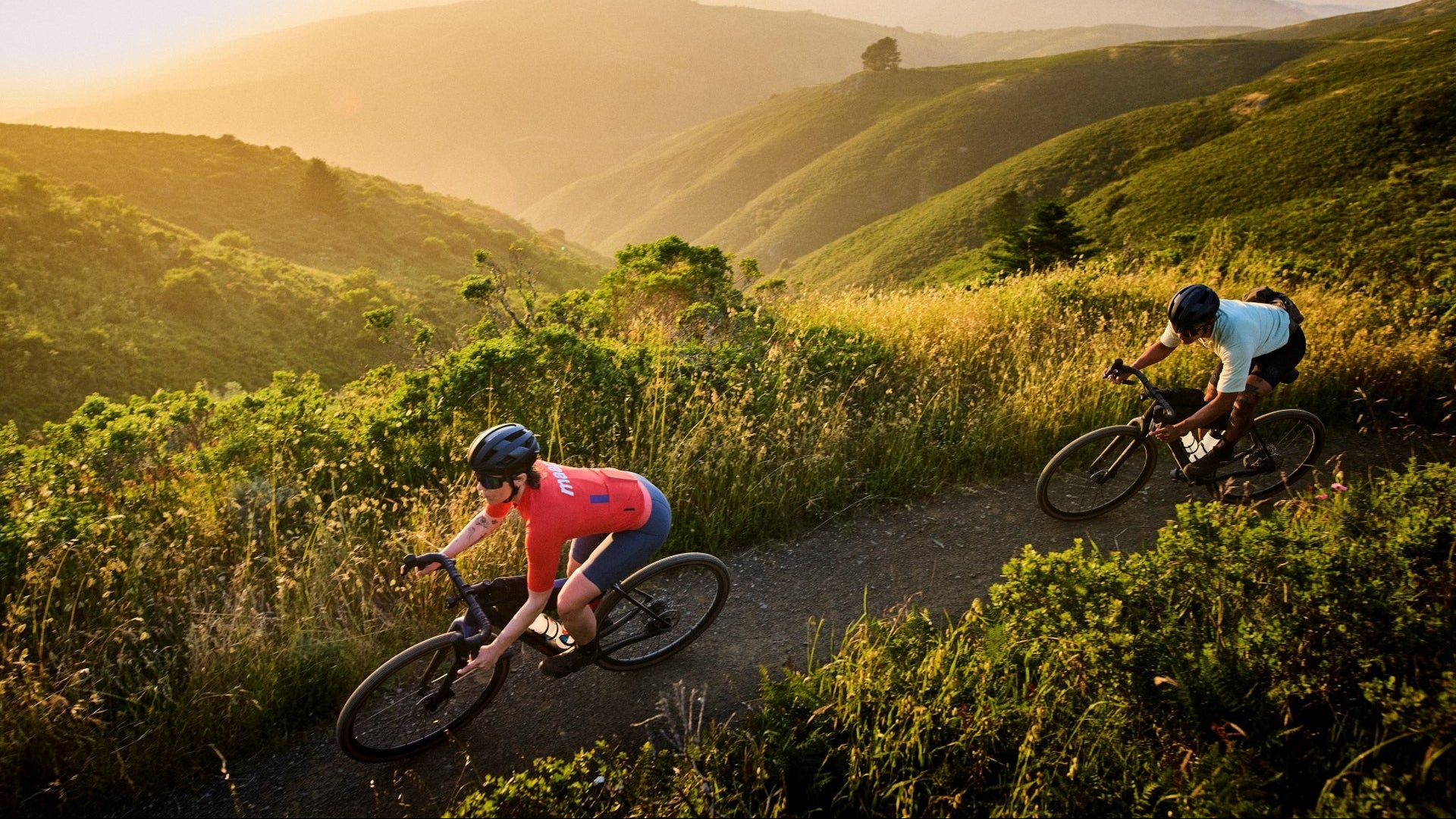 Two cyclists riding on a trail with mountains in the background during sunset.