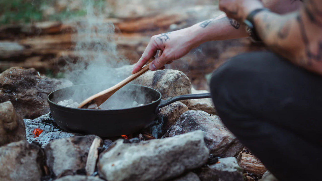 Person cooking over a campfire using a pan and wooden spoon on rocks.