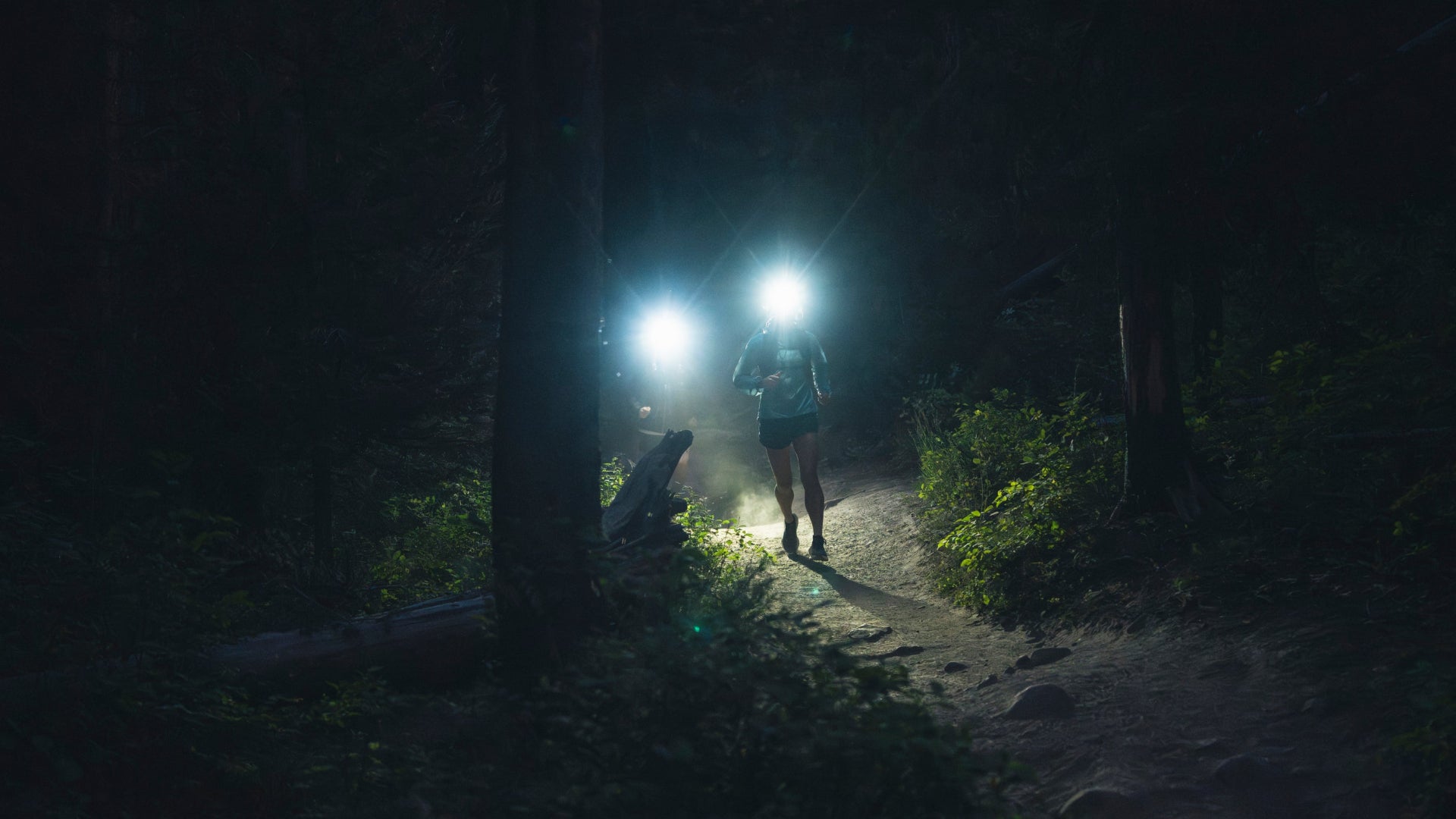 Two people running on a forest path at night with headlamps illuminating the way.