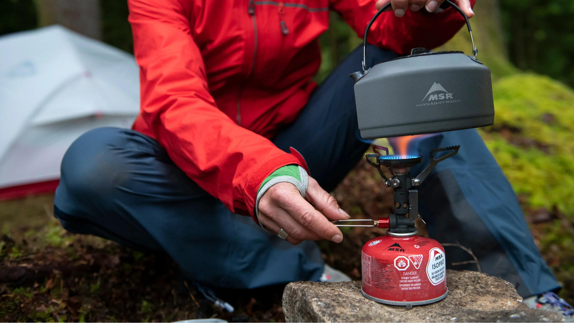Person in red jacket using a camping stove in a forest setting.