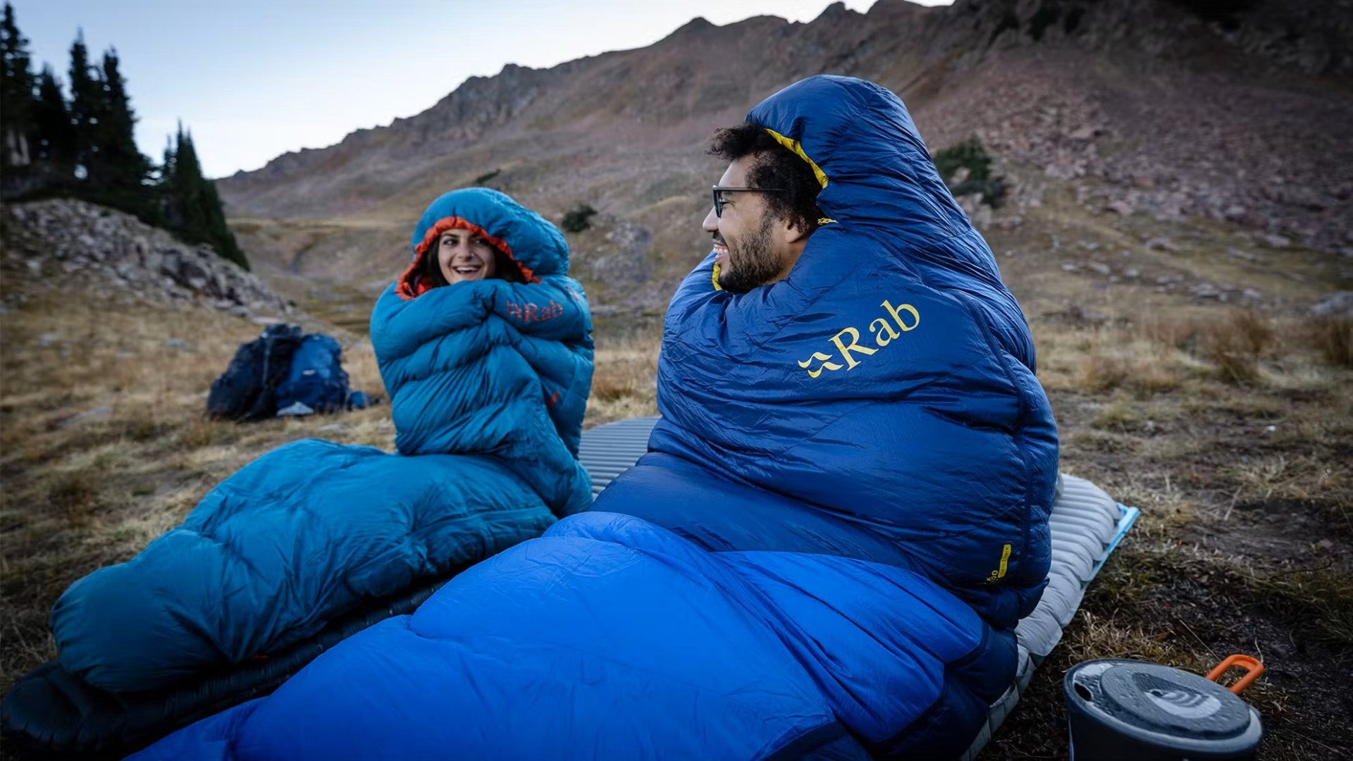 Two people in blue sleeping bags in a mountainous outdoor setting