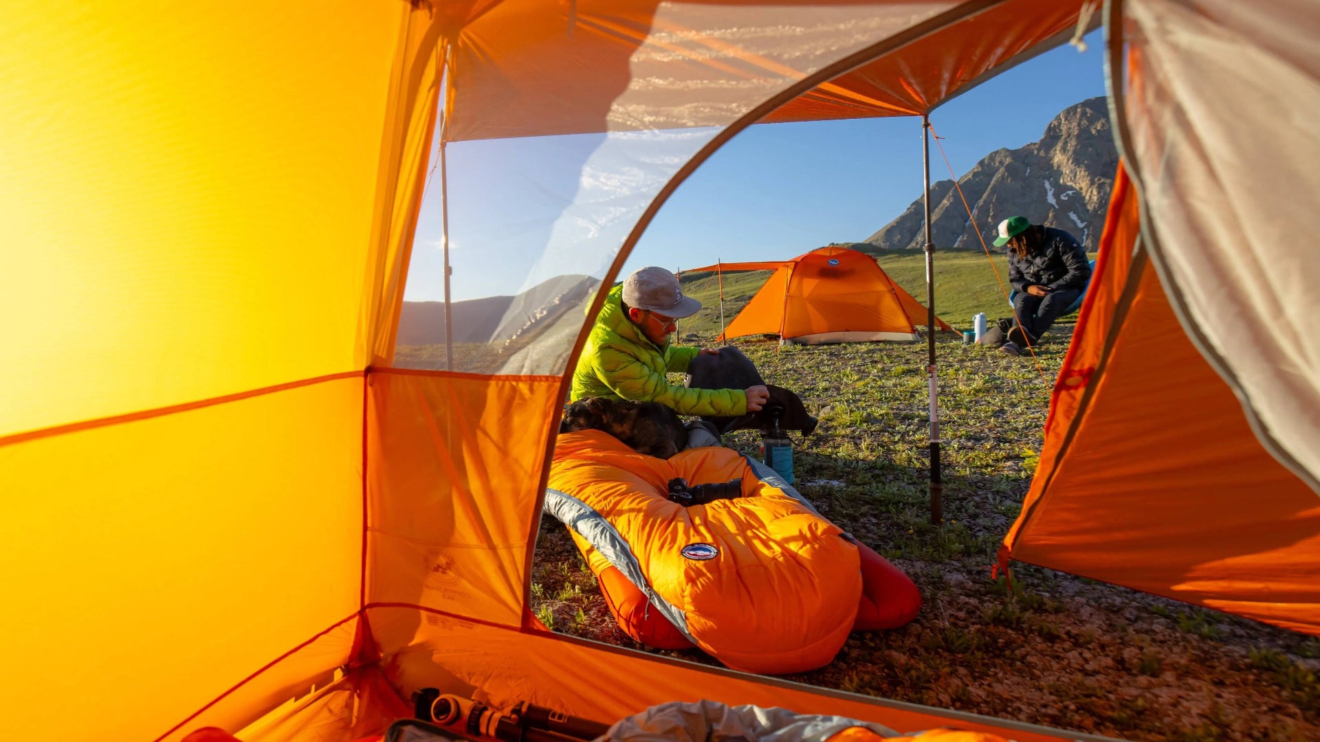 Person setting up a tent in a mountainous area with another tent visible in the background.