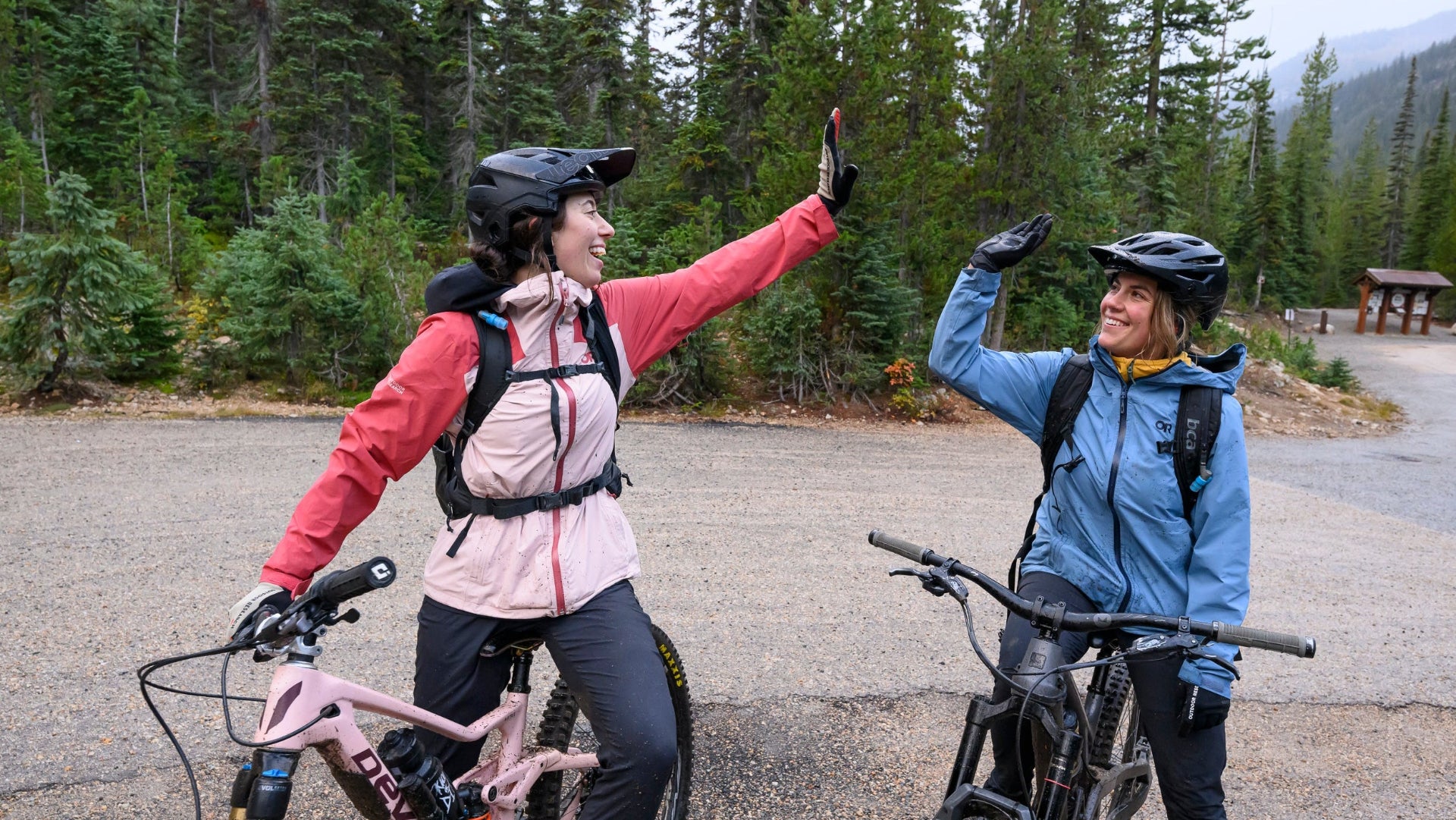 Two cyclists giving high-fives on a trail with trees and mountains in the background